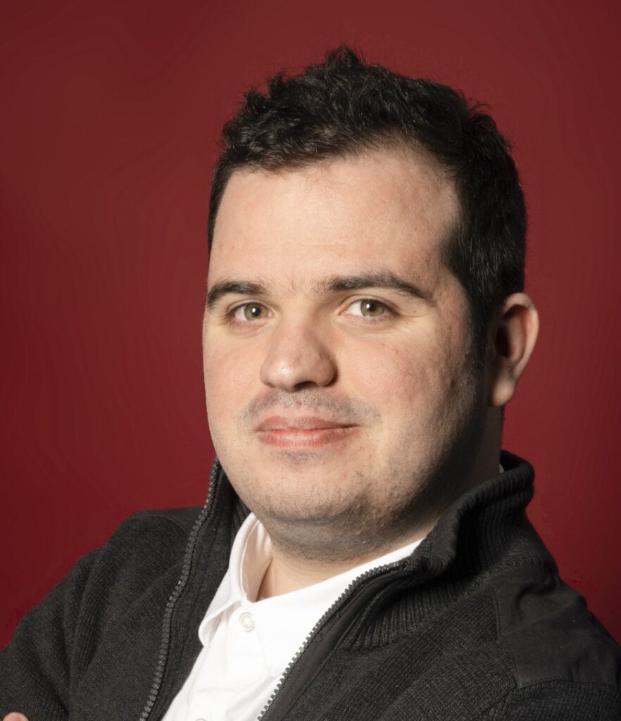Head-and-shoulders portrait of a man with short dark hair, wearing a white shirt and dark zip-up jacket against a solid red background.